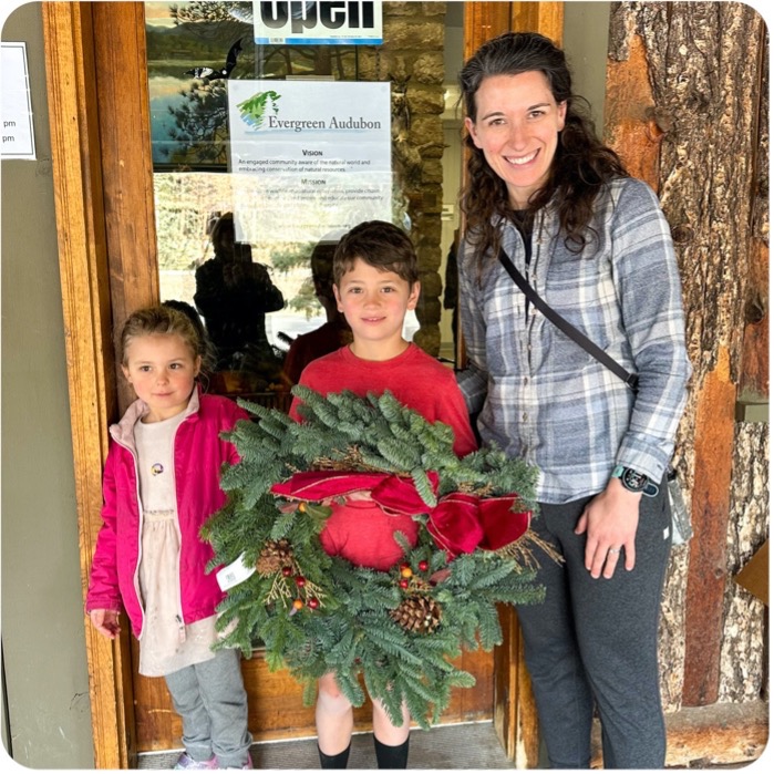 two children, the one in the middle holding a wreath, and a mom in front of the nature center door